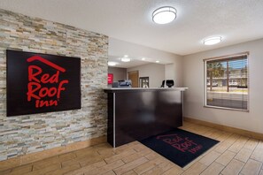A Red Roof Inn reception desk with a stone wall backdrop, a wooden floor, and a window with blinds. - Red Roof Inn Columbia, SC Airport (West Columbia)