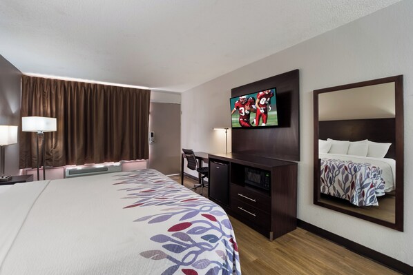 A hotel room with a large bed, a flat-screen TV mounted on the wall, a desk with a chair, a microwave, and a refrigerator.