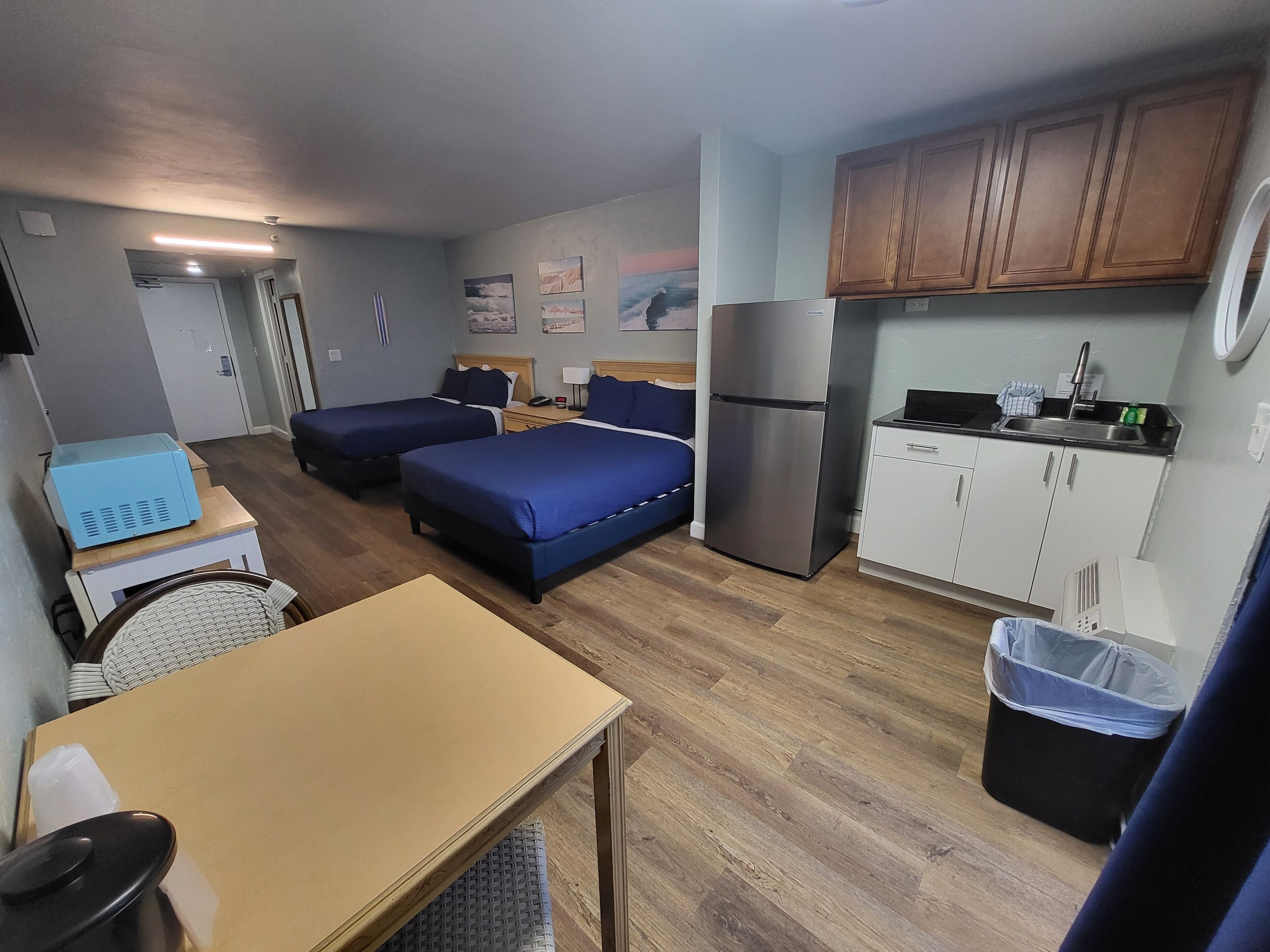 Quarto de hotel compacto com duas camas, uma kitchenette, uma pequena mesa de jantar e uma lixeira.