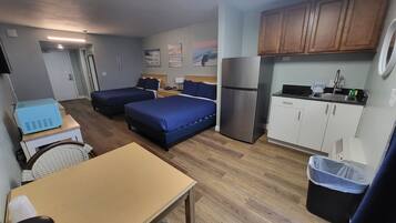 Quarto de hotel compacto com duas camas, uma kitchenette, uma pequena mesa de jantar e uma lixeira.