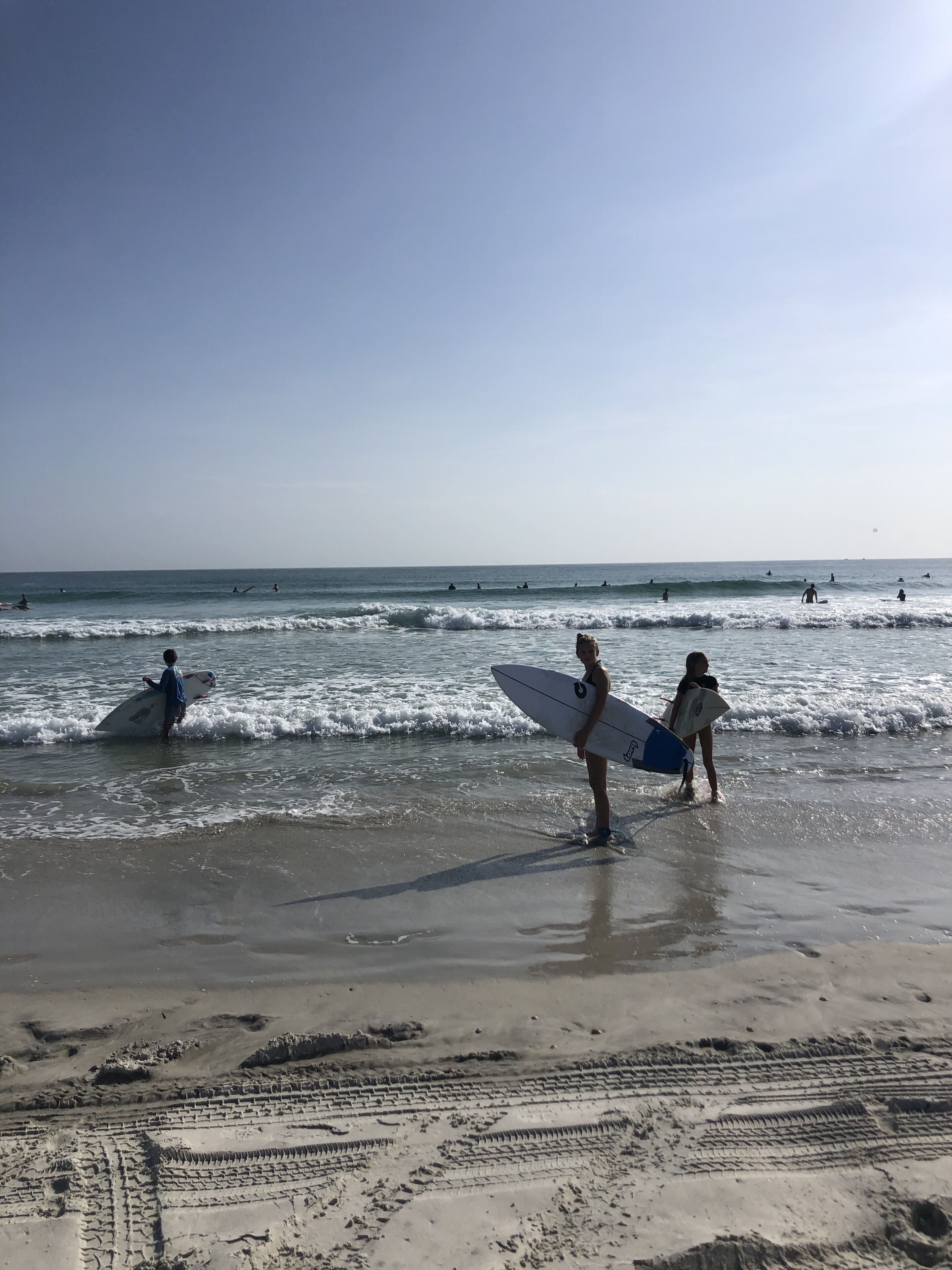 Une plage où l’on voit des gens tenant des planches de surf, marchant le long du rivage, et d’autres faisant de la planche à voile dans l’eau.
