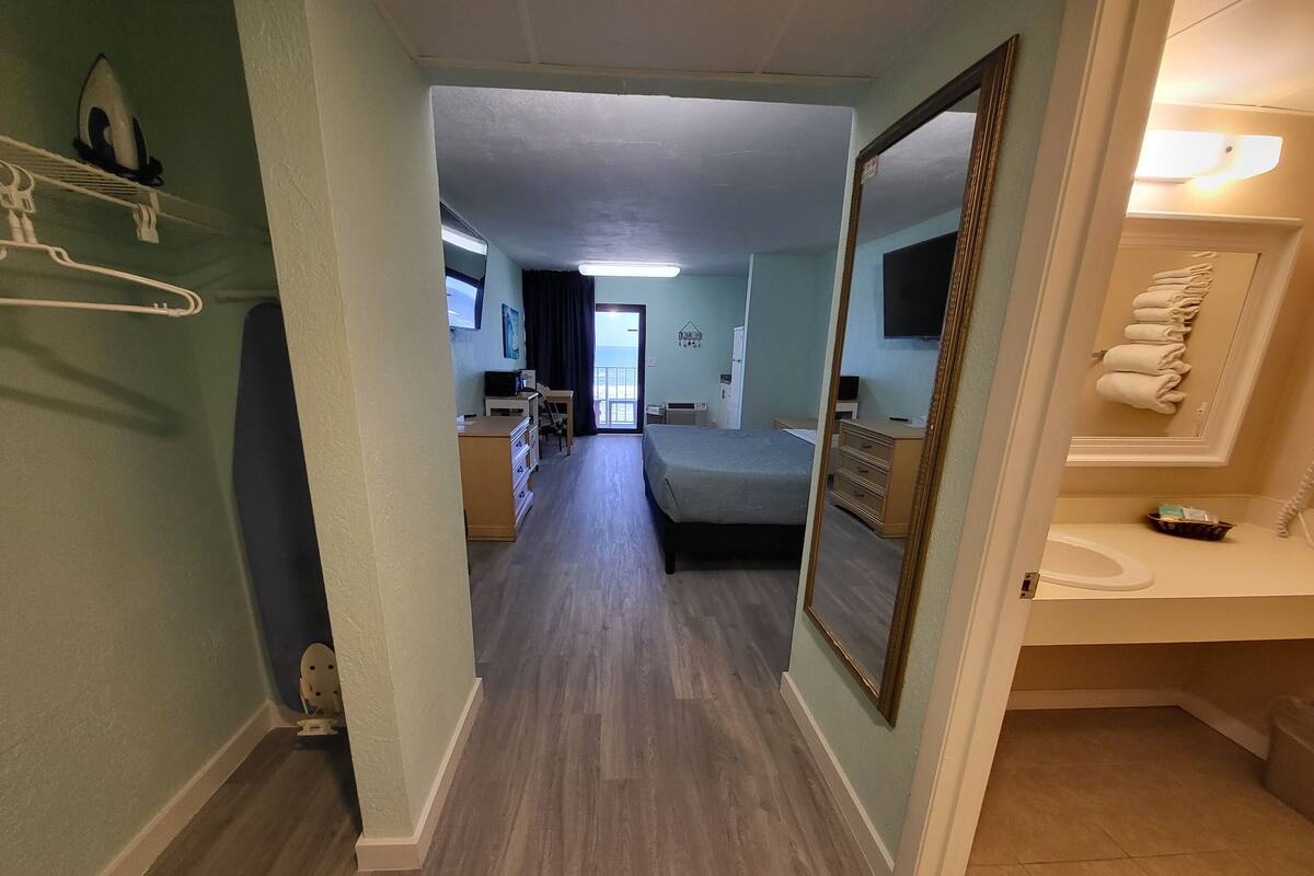 A hotel room with a bed, a desk, a chair, a dresser, a mirror, and a bathroom with a sink and towel rack.