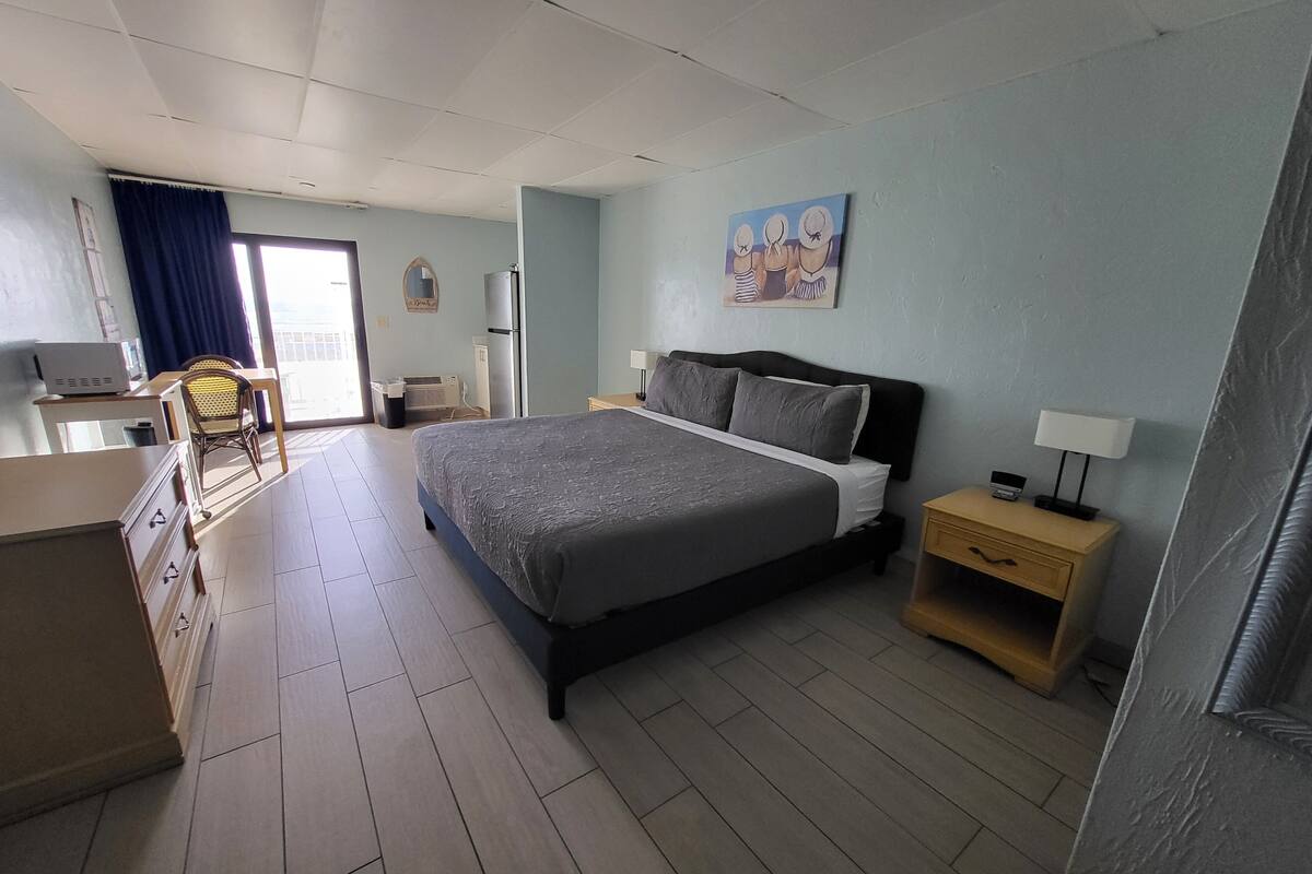 A bedroom with a bed, a desk, a chair, a nightstand, and a painting on the wall.