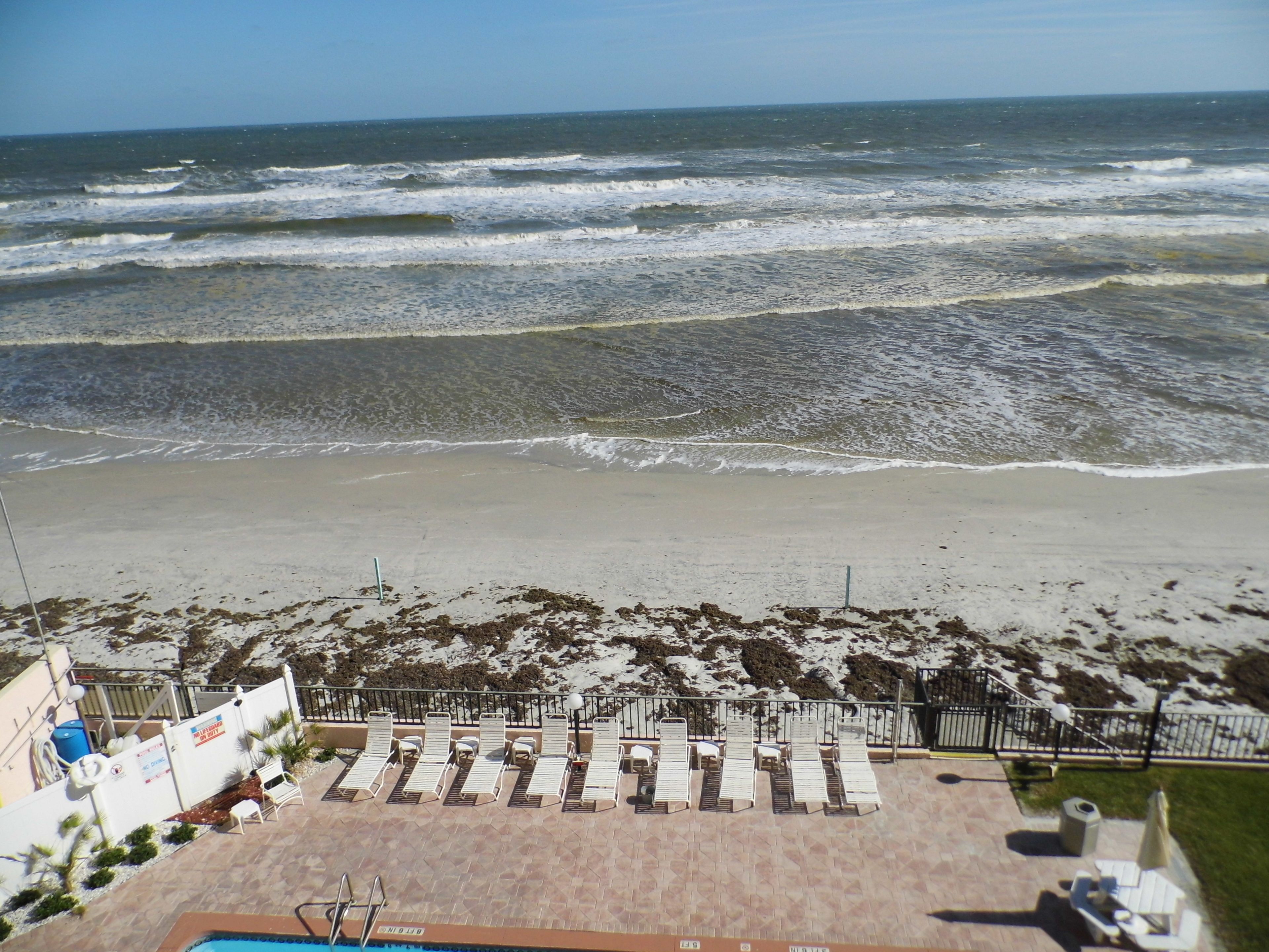 A beach view with a pool area, sun loungers, and a fence.