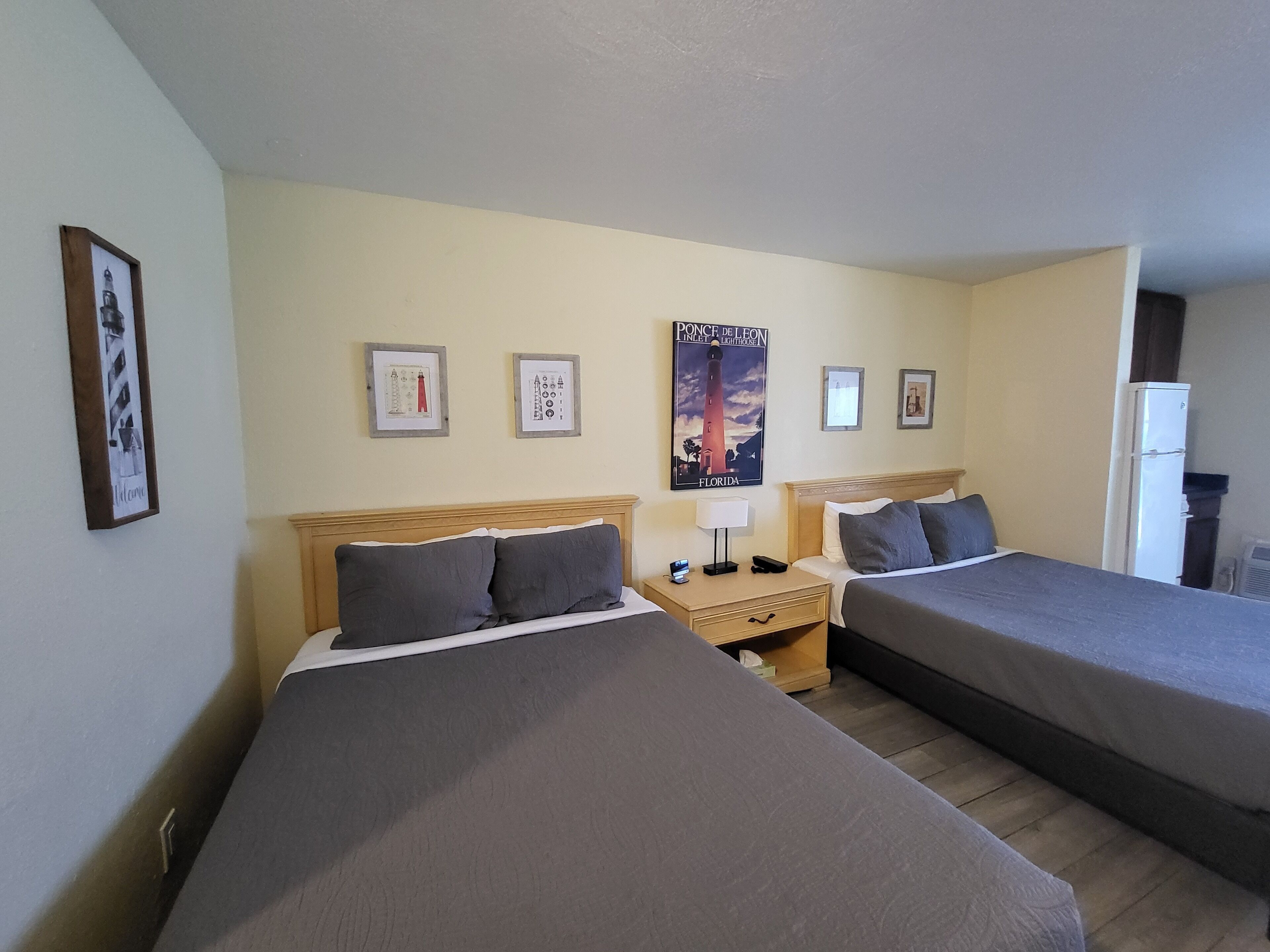A hotel room with two single beds, a nightstand with a lamp, a poster on the wall, and a refrigerator.