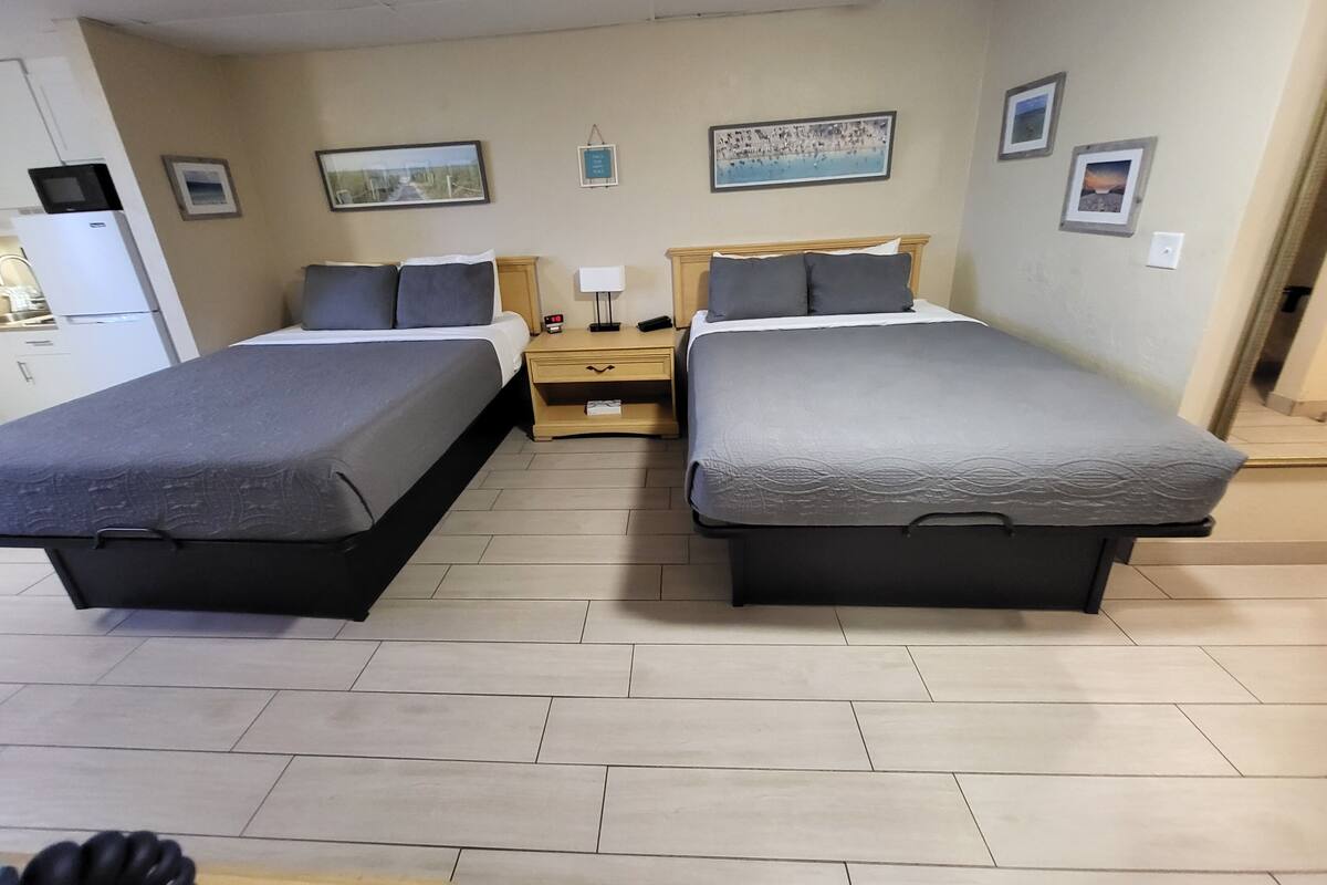 A hotel room with two beds, a small desk, a nightstand, and a refrigerator.