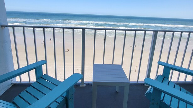 A balcony with two blue Adirondack chairs and a small table, overlooking a sandy beach and the ocean.