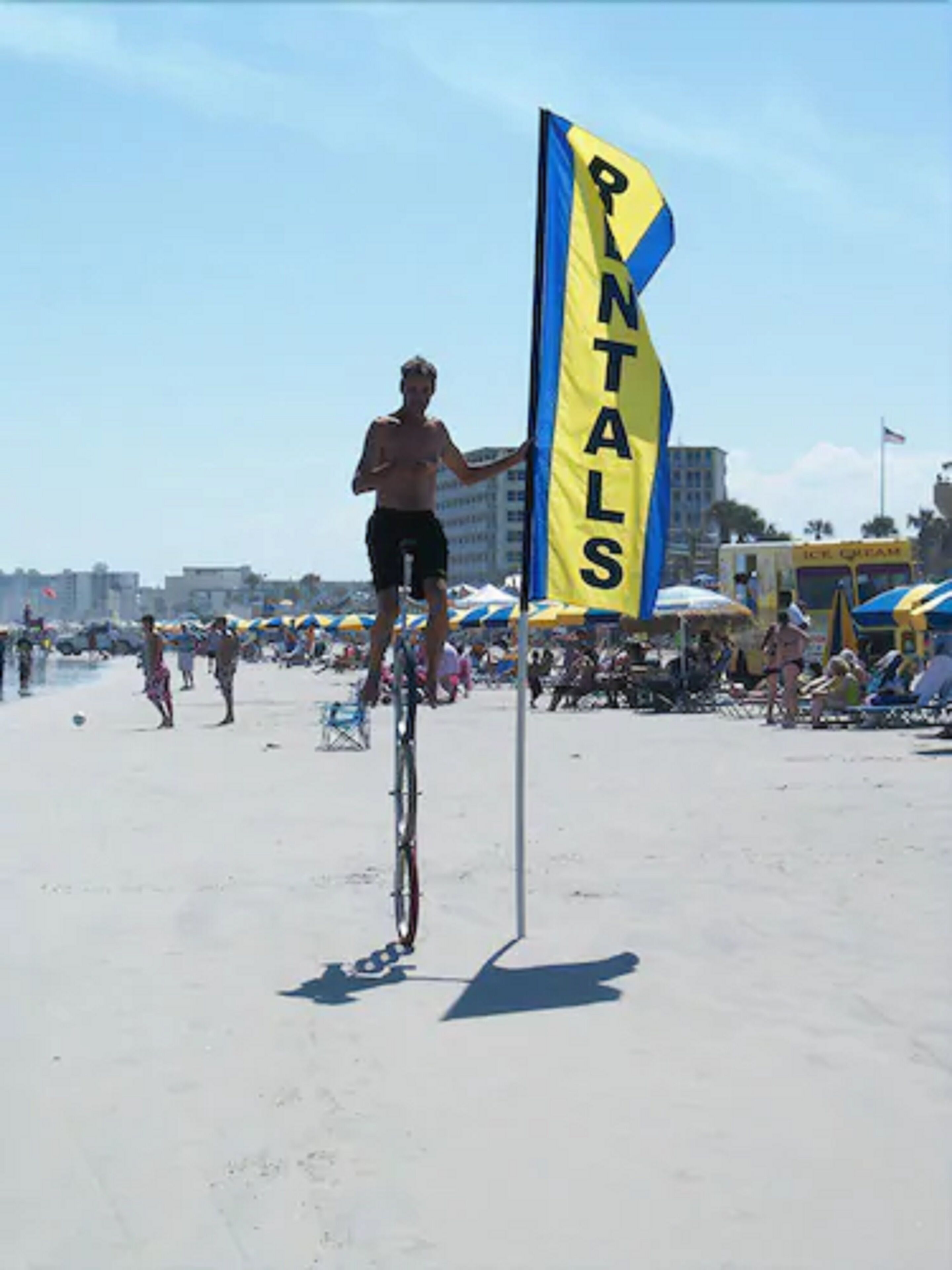 Eine Person fährt auf einem Einrad über einen sandigen Strand, mit einer Flagge mit dem Aufdruck 'PINTALS'.