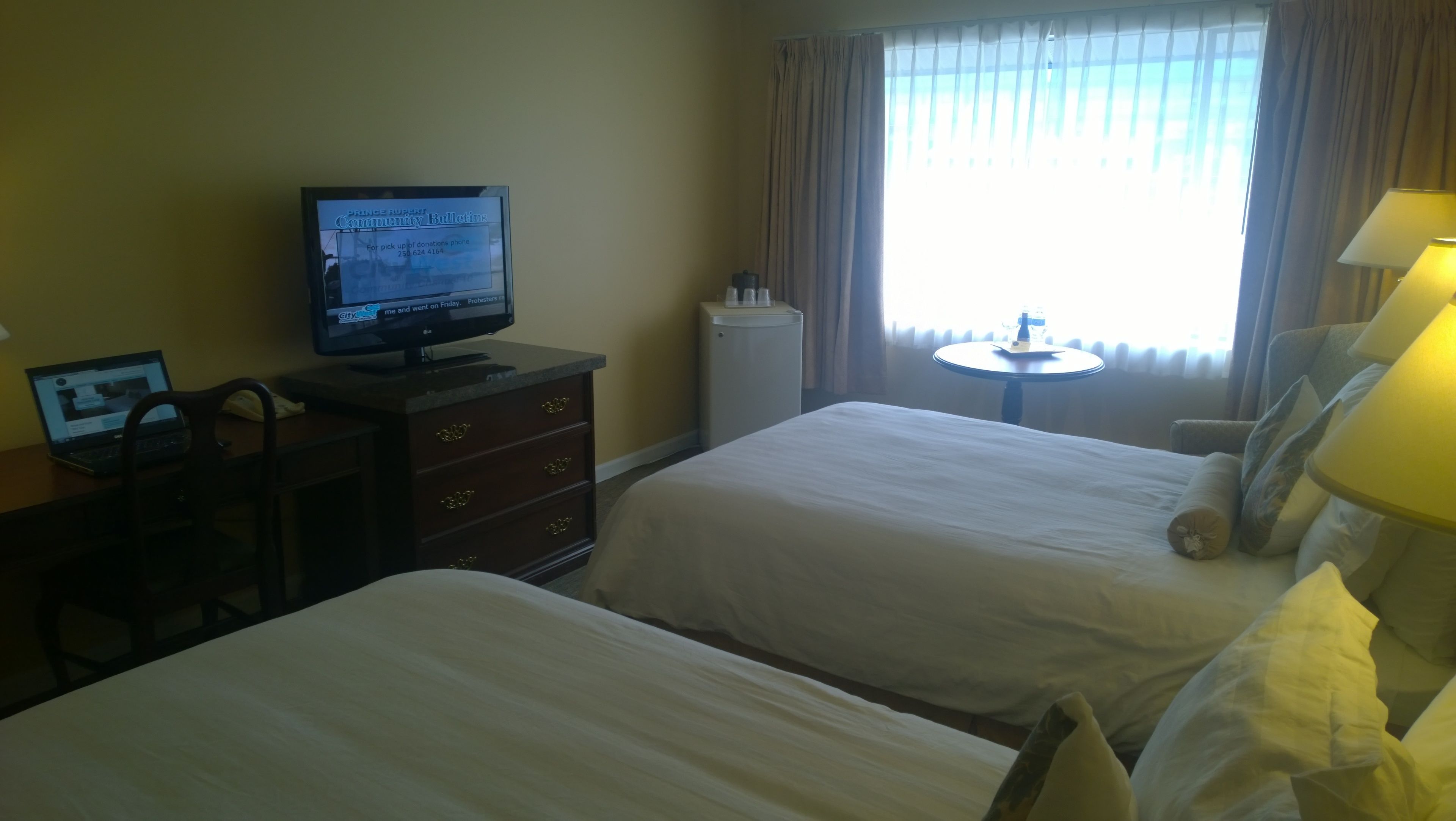 A hotel room with two beds, a desk with a laptop, a television, a small round table with a lamp, and a window with curtains.