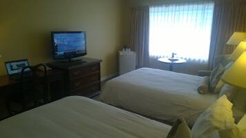 Une chambre d’hôtel avec deux lits, un bureau avec un ordinateur portable, une télévision, une petite table ronde avec une lampe et une fenêtre avec des rideaux.