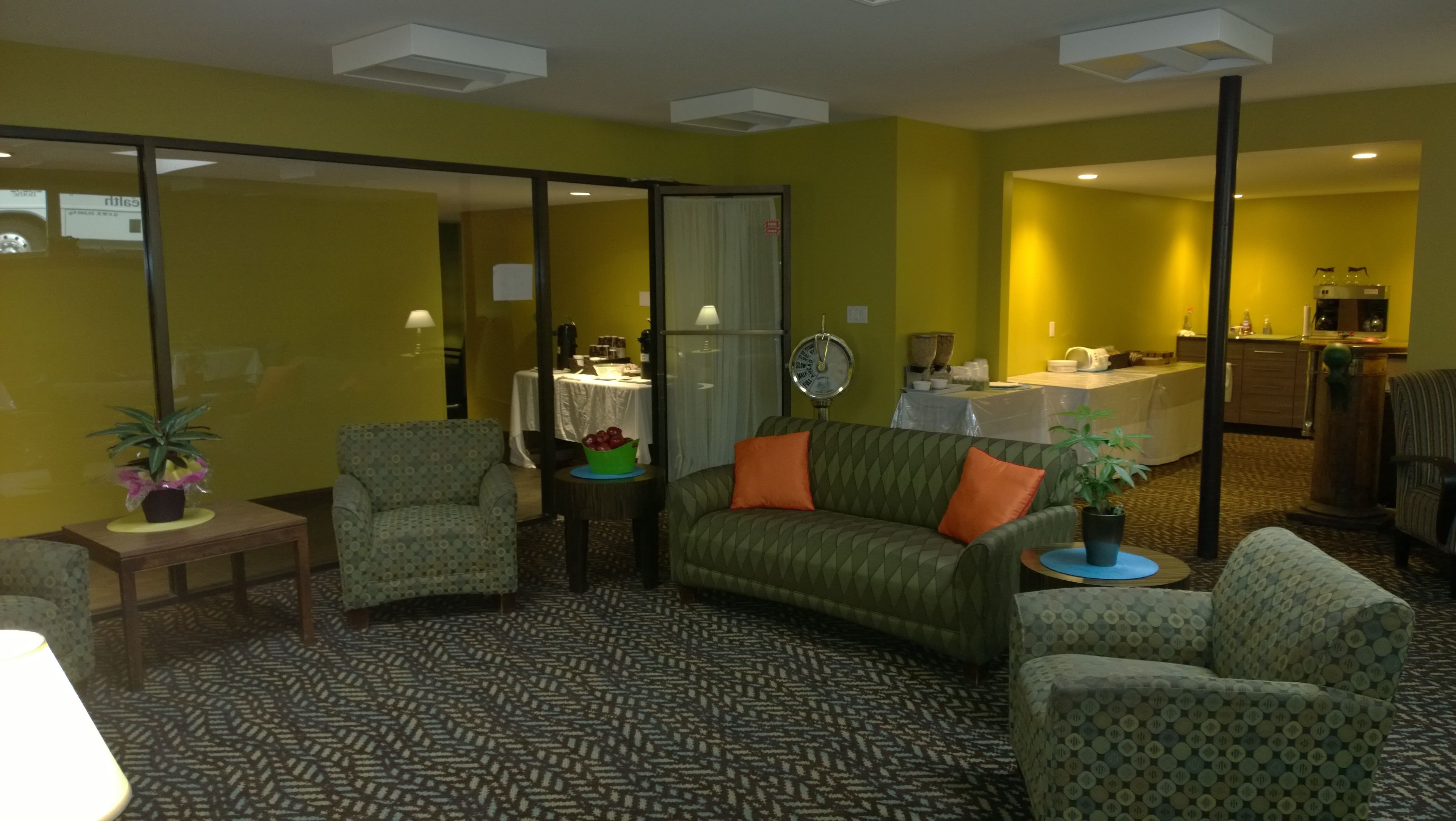 A room with a patterned carpet, a sofa with orange cushions, a chair, a small table with a lamp, and a potted plant.