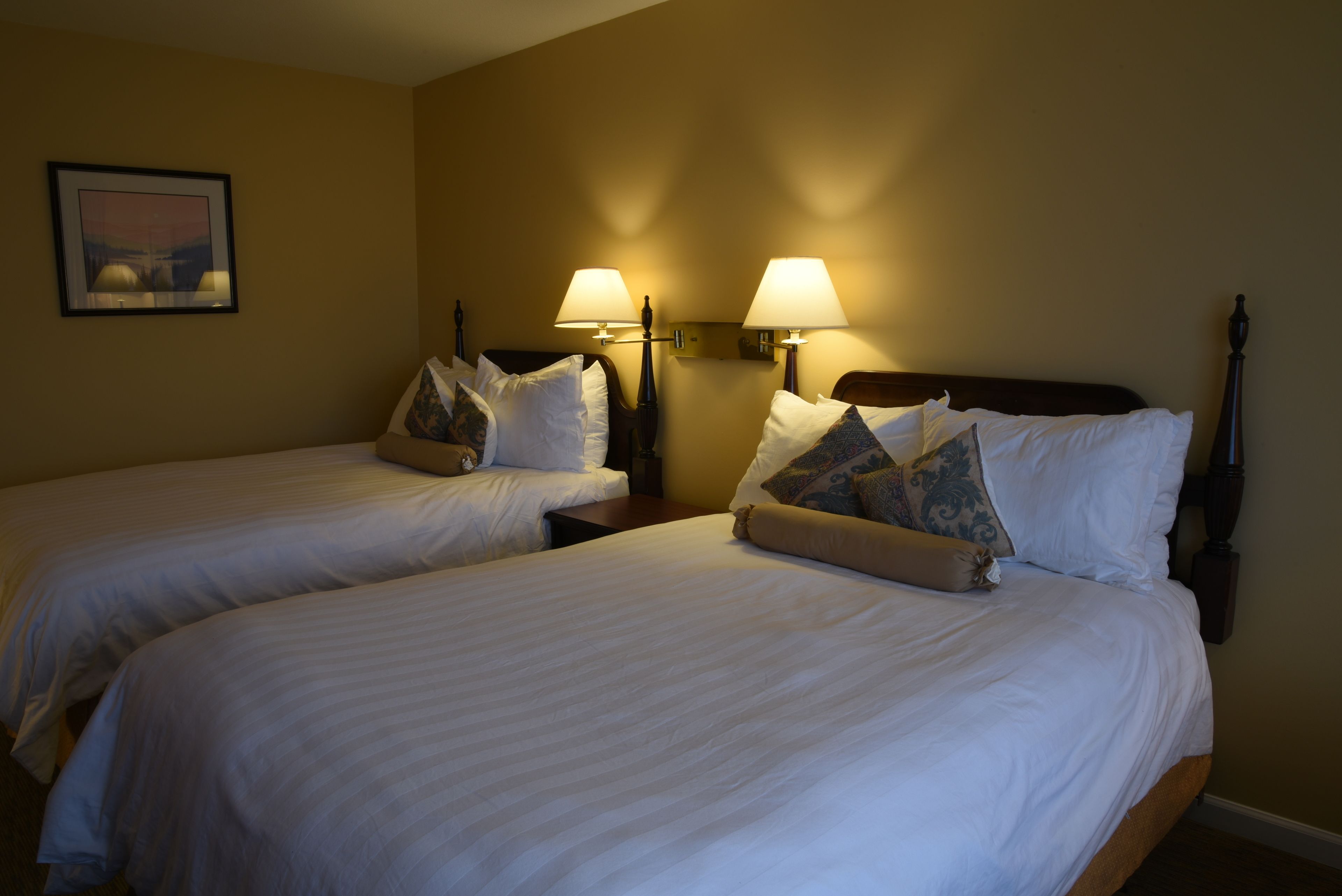 A hotel room with two beds, each with white linens and decorative pillows. There are wall-mounted lamps on either side of the beds.