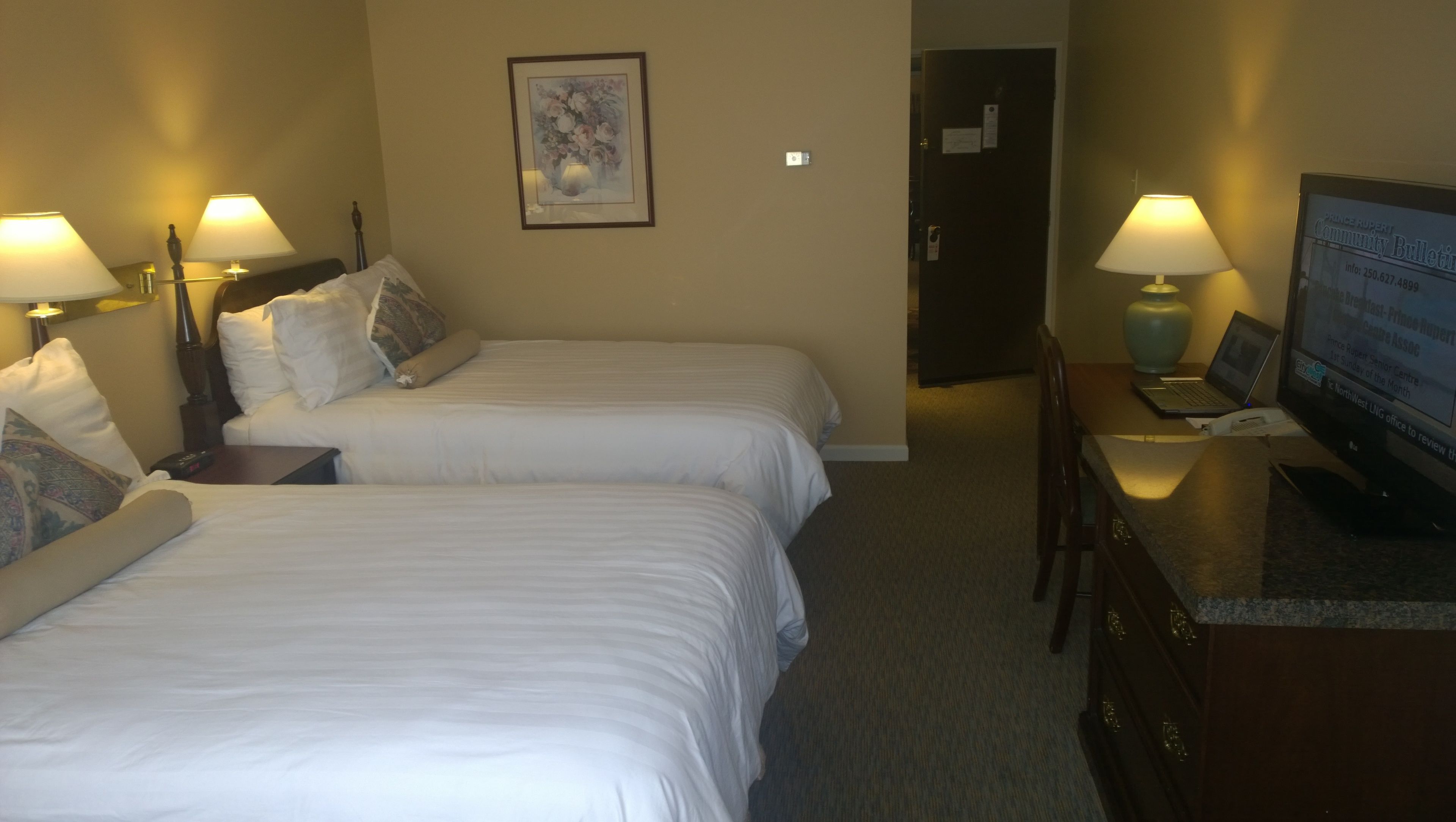 Ein Hotelzimmer mit zwei Betten, einem Schreibtisch mit Computer und einem Fernseher, der einen Informationsaushang zeigt.