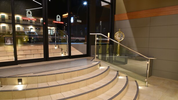 Un escalier incurvé et illuminé mène à l’entrée d’un bâtiment, qui est dotée de portes en verre et d’une horloge.