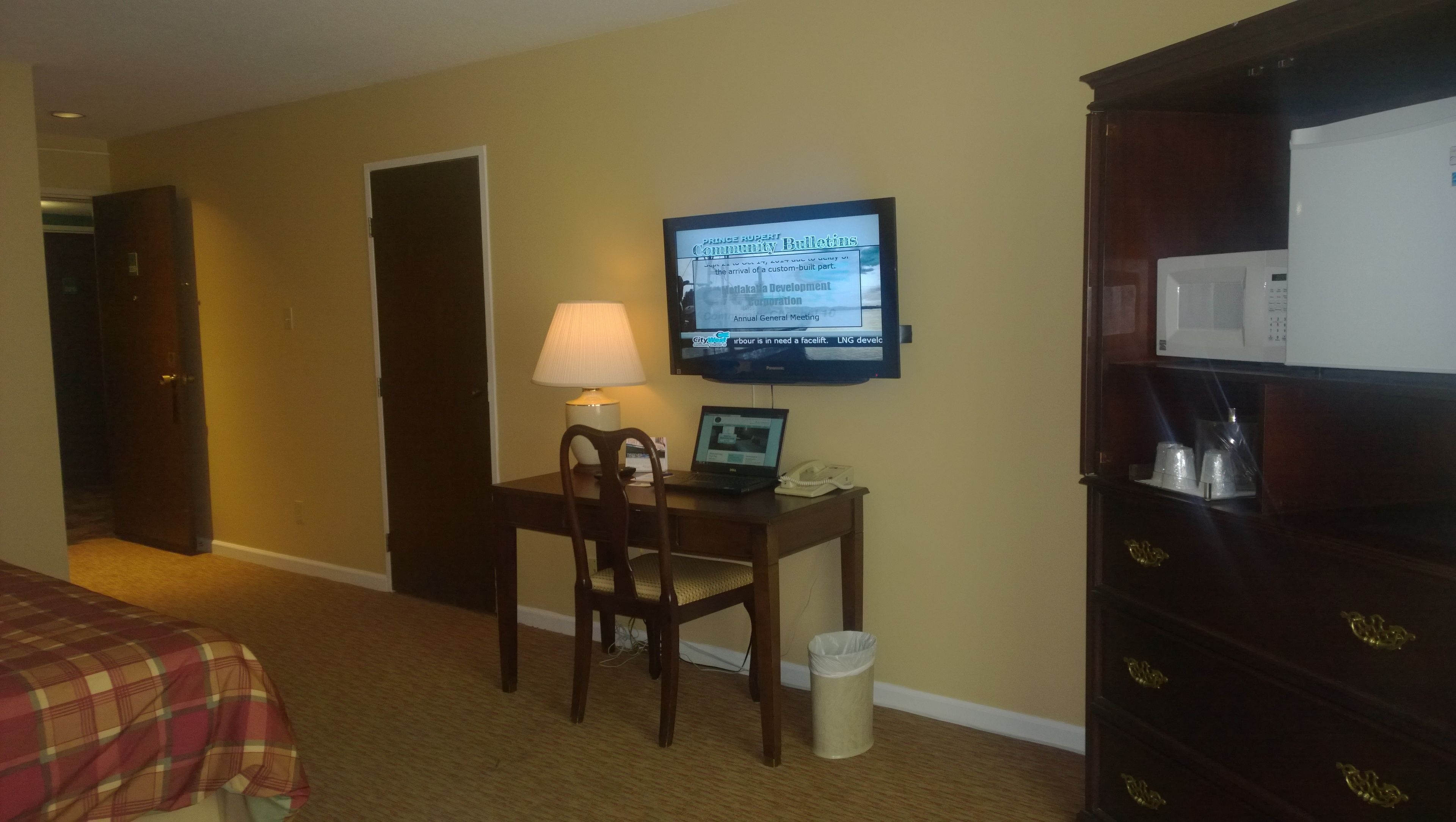 Une chambre d’hôtel comprenant un lit, un bureau avec un ordinateur, un téléviseur diffusant des informations locales, une lampe et une commode.