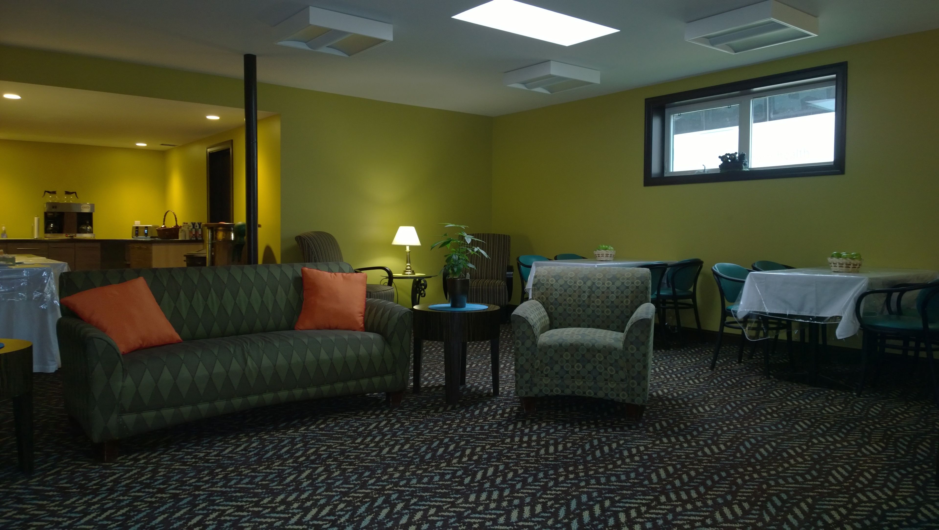 A room with a green sofa, a patterned carpet, a dining table with chairs, and a window with blinds.