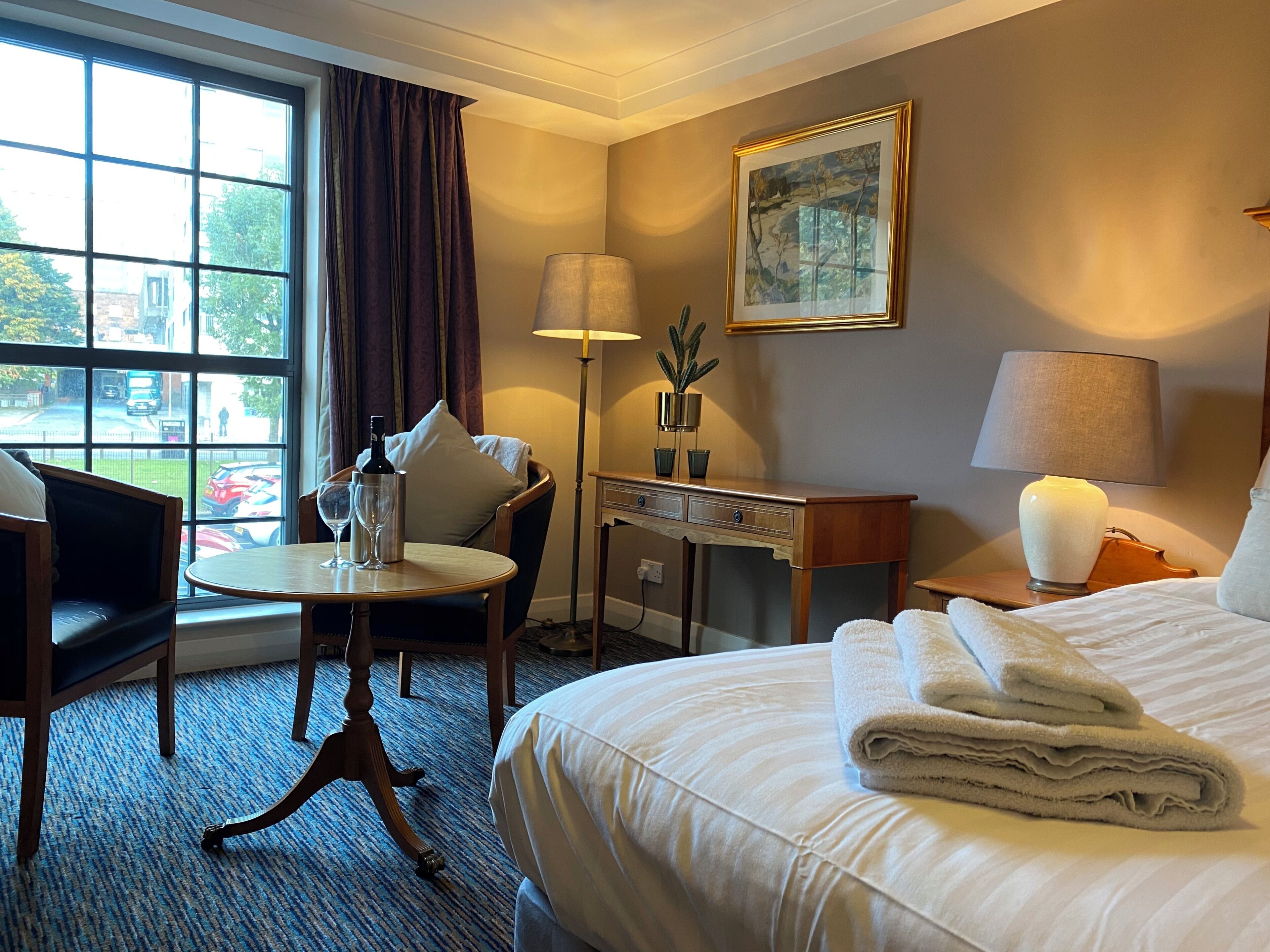 A hotel room with a bed, a desk, a chair, a table, a lamp, a painting, and a window with curtains.