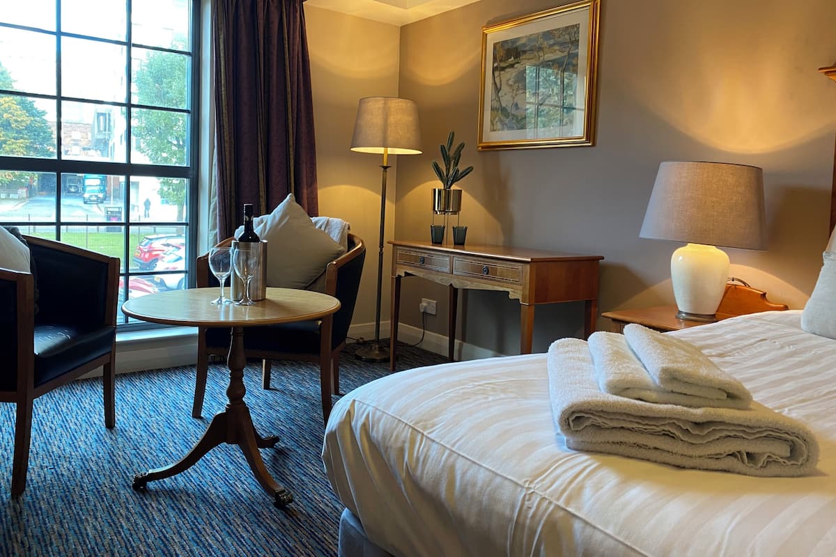 A hotel room with a bed, a desk, a chair, a table, a lamp, a painting, and a window with curtains.