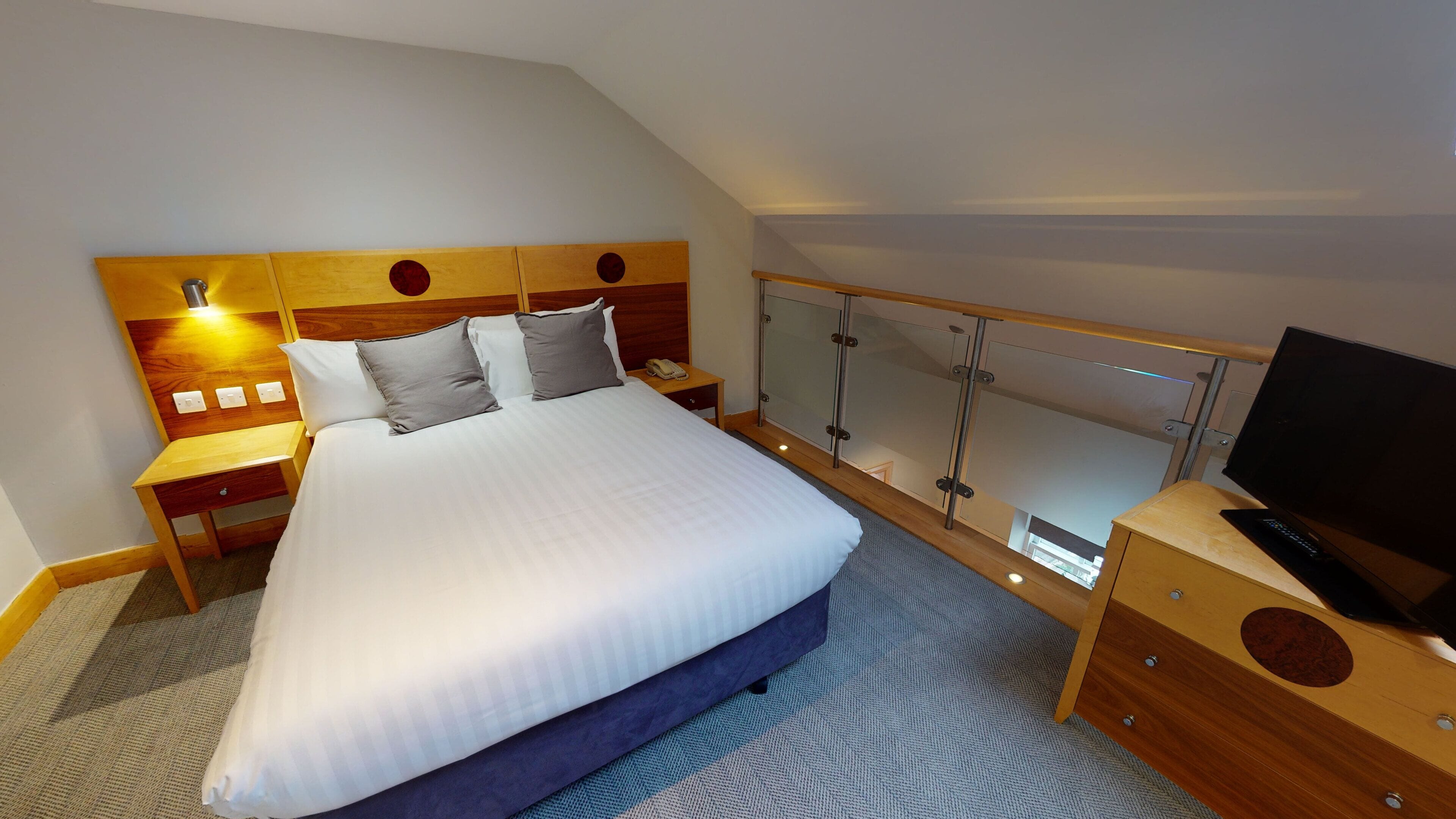 Habitación de hotel con una cama grande, cabecera de madera, mesitas de noche, televisor y balcón con barandilla de vidrio.