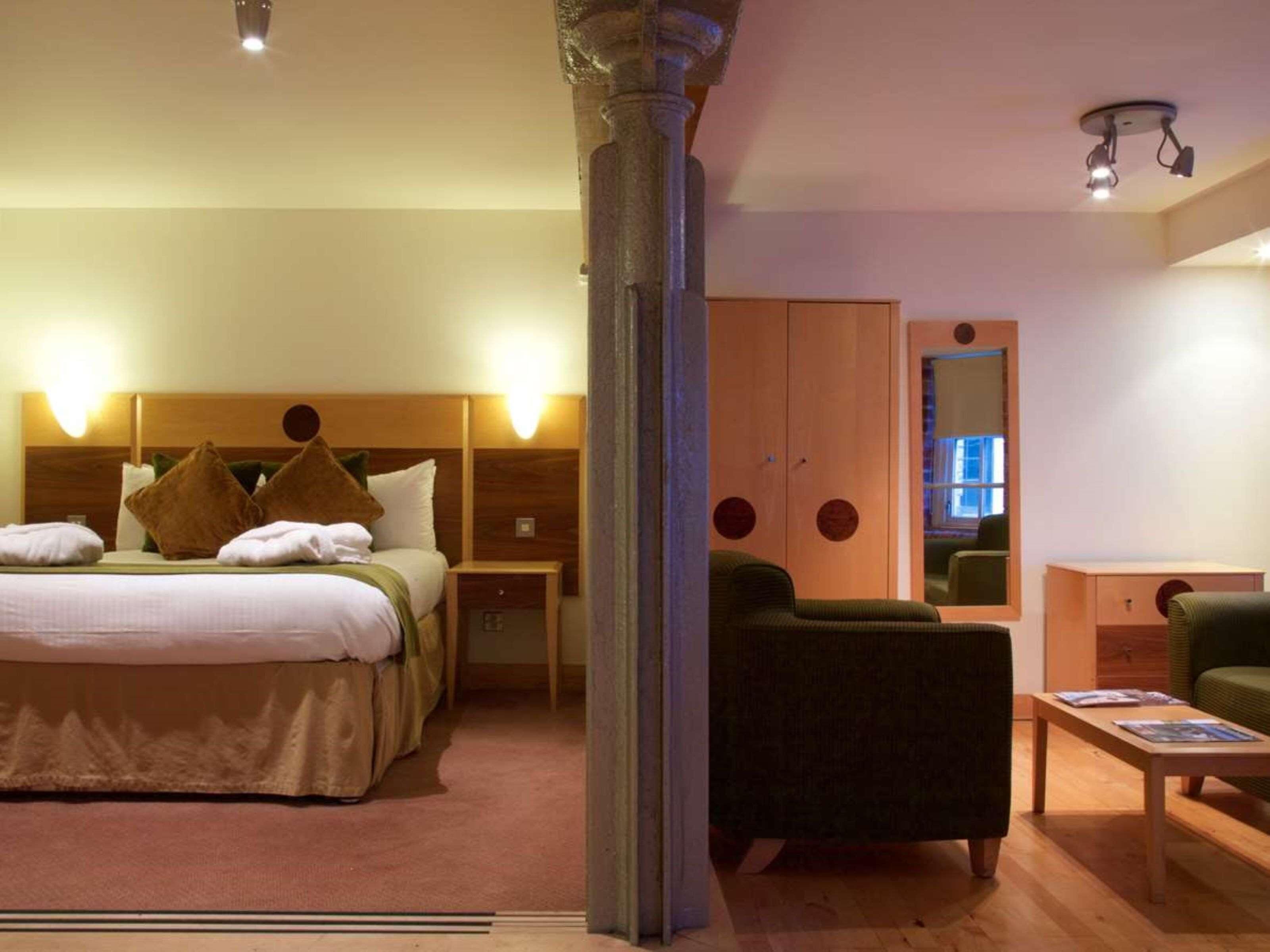 A hotel room with a bed, bedside tables, a green armchair, a wooden wardrobe, and a mirror.