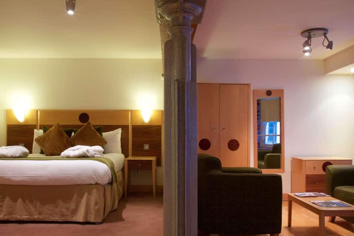 A hotel room with a bed, bedside tables, a green armchair, a wooden wardrobe, and a mirror.