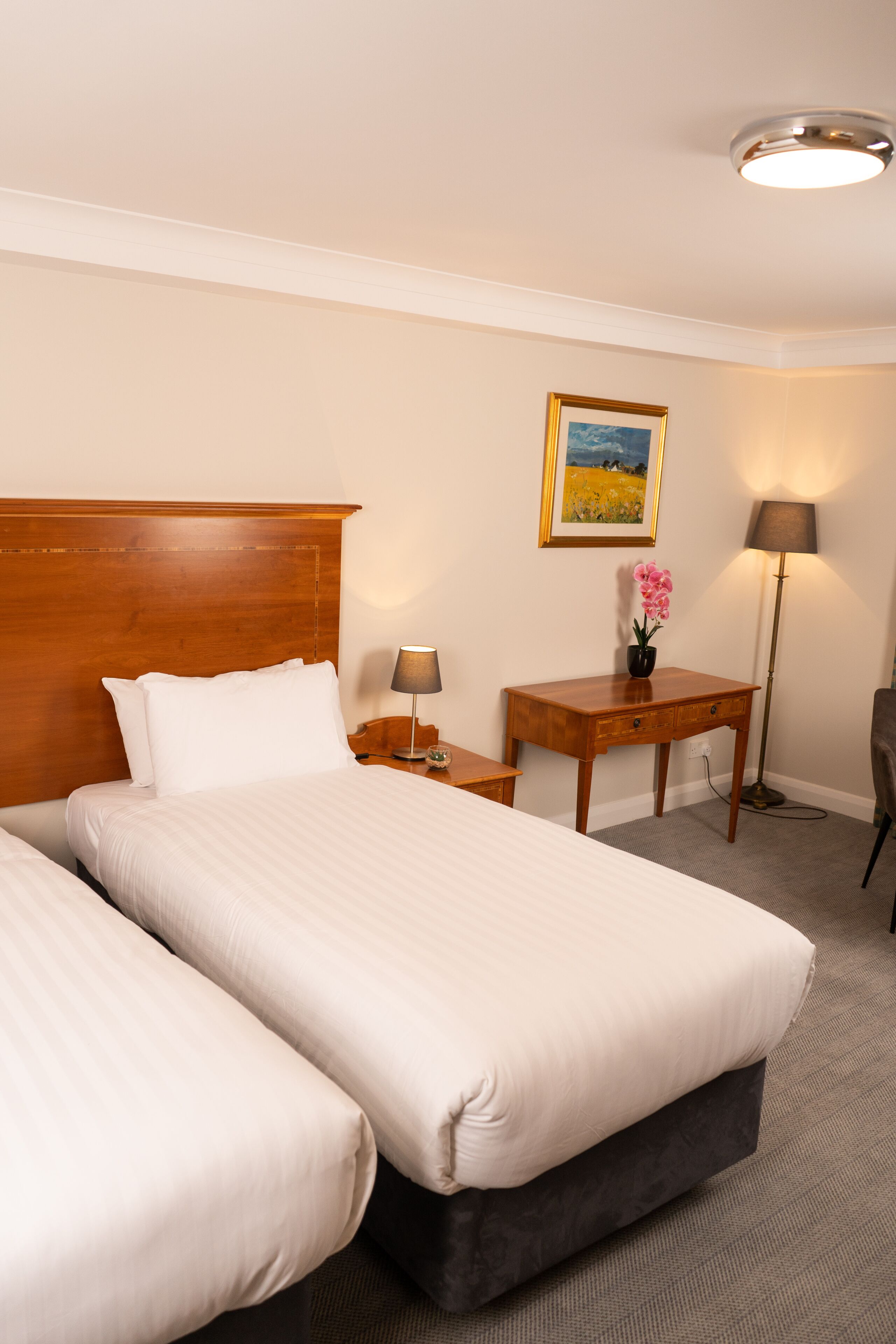 Hotelkamer met twee bedden, een houten hoofdeinde, een nachtkastje met een lamp, een tafeltje met een bloevas en een ingelijst schilderij aan de muur.