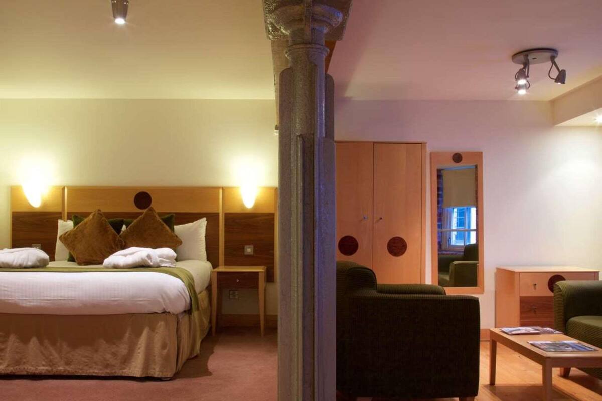 A hotel room with a bed, bedside tables, a green armchair, a wooden wardrobe, and a small table with magazines.