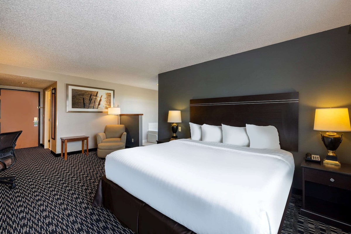 A hotel room with a large bed, two bedside tables with lamps, a chair, a desk with a laptop, and a framed picture on the wall.