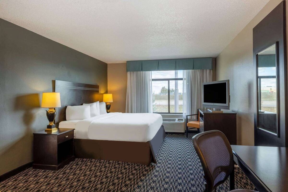 A hotel room with a bed, two bedside lamps, a desk with a television, a chair, and a large window with curtains.