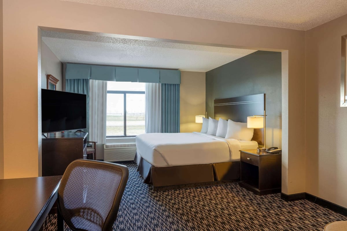 A hotel room with a large bed, a desk with a chair, a TV, a nightstand with a lamp, and a window with curtains.