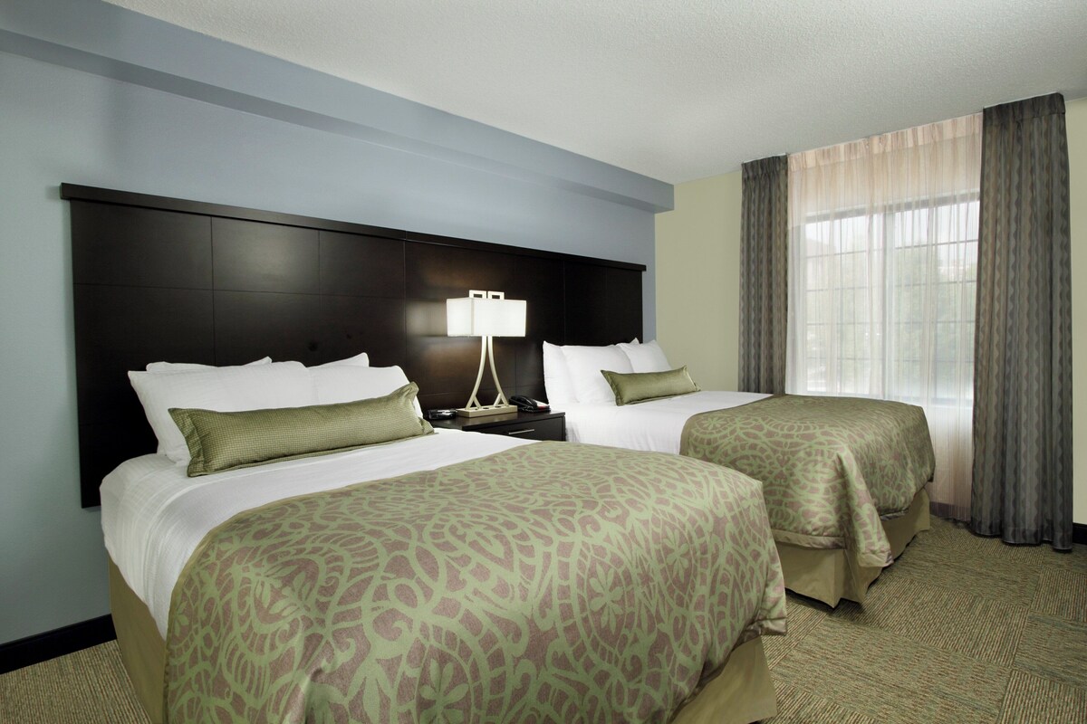 A hotel room with two beds, a nightstand, a lamp, and a window with curtains.