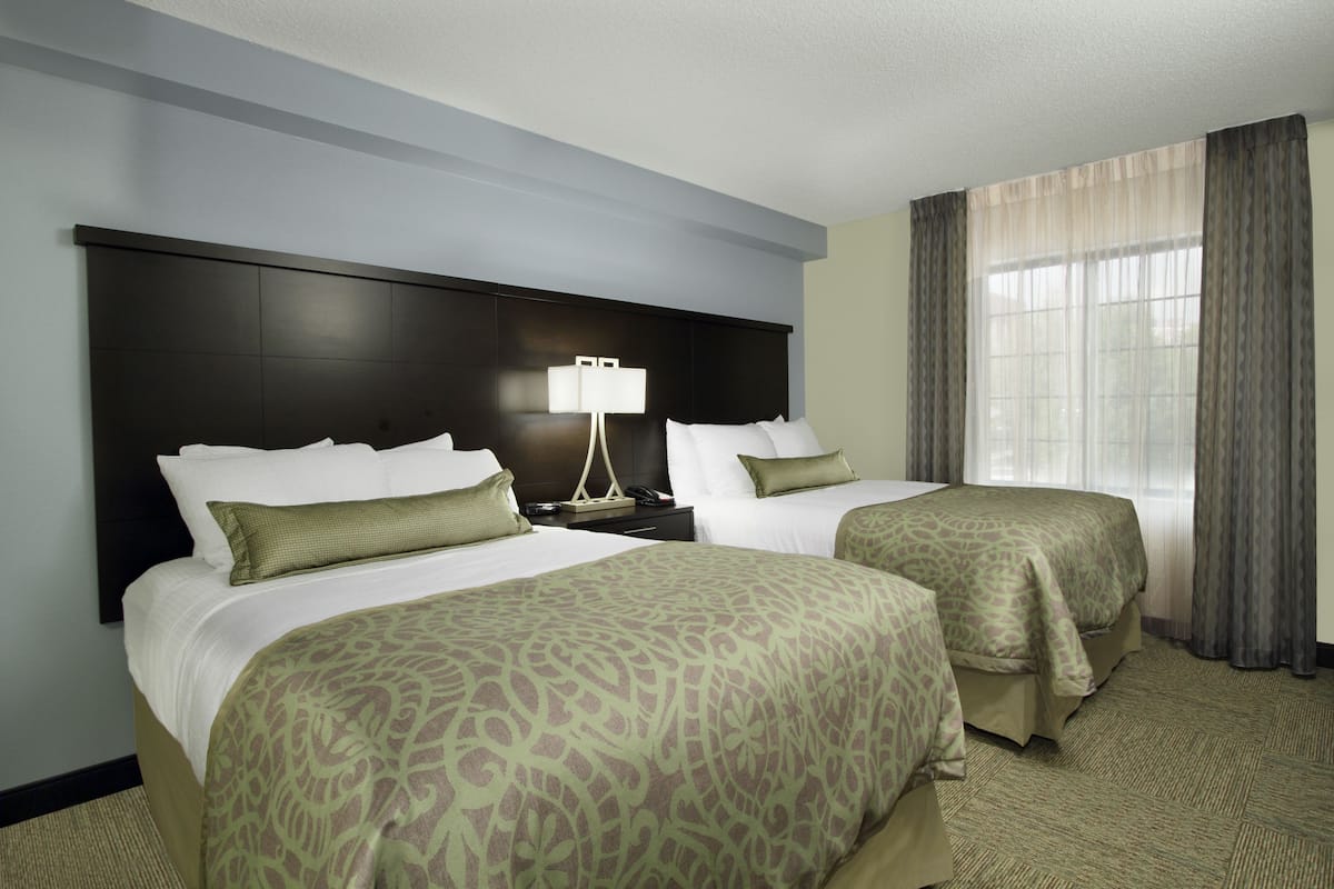 A hotel room with two beds, a nightstand, a lamp, and a window with curtains.