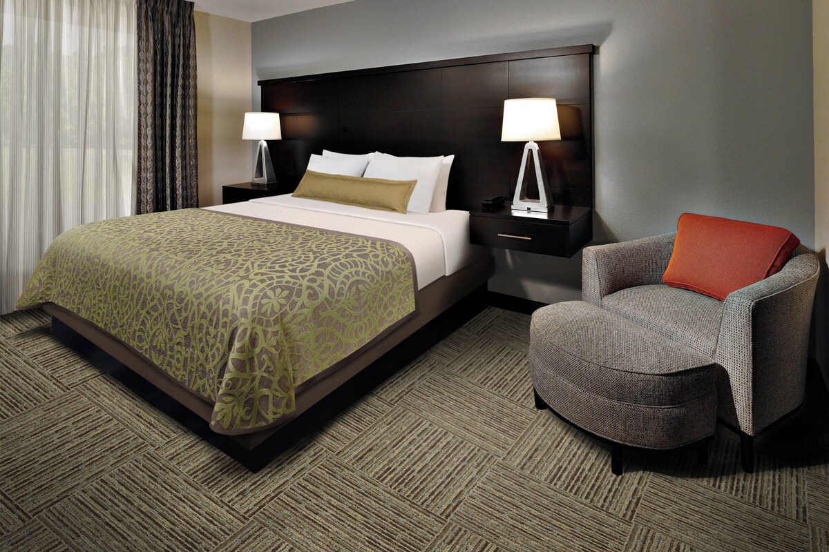A hotel room with a large bed, two bedside tables with lamps, a chair, and a carpeted floor.