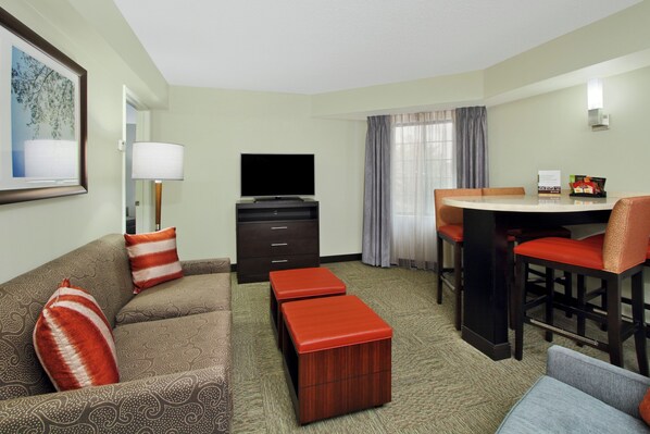 A hotel room with a sofa, a TV, a desk with chairs, and a small table.