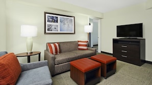 A hotel room with a grey sofa, a blue armchair, two wooden ottomans, a flat-screen TV, a dark wooden cabinet, and a framed picture on the wall. - Staybridge Suites Tysons - McLean by IHG (McLean)