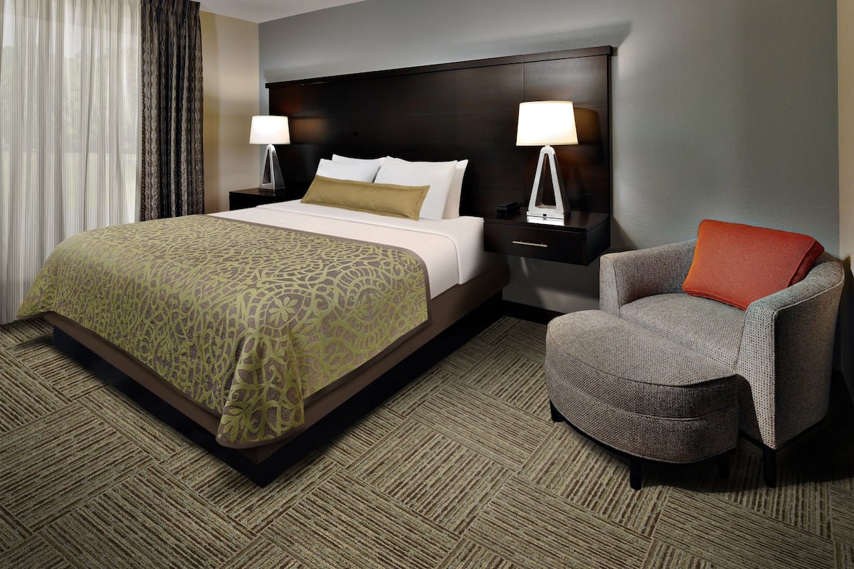 A hotel room with a large bed, two bedside tables with lamps, a grey armchair, and a patterned carpet.