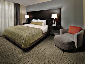 A hotel room with a large bed, two bedside tables with lamps, a grey armchair, and a patterned carpet. - Staybridge Suites Tysons - McLean by IHG (McLean)
