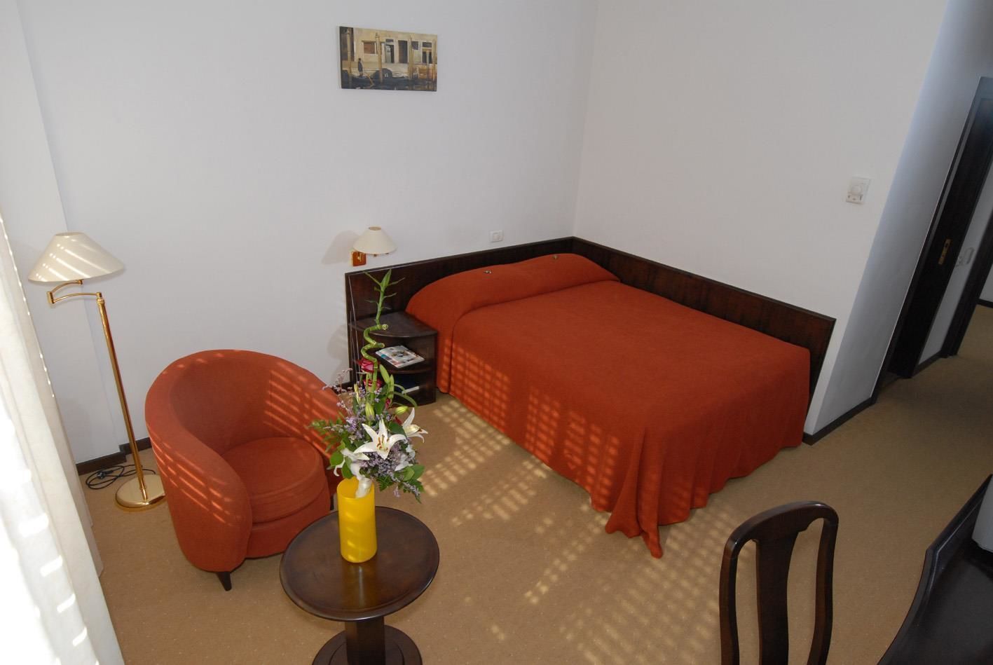 Habitación de hotel con una cama individual, una silla, una mesita con un jarrón de flores, una lámpara y un cuadro en la pared.