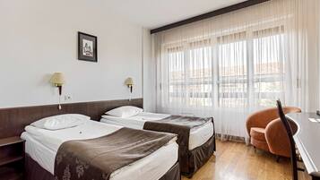 A hotel room with two single beds, a desk, a chair, and a large window with sheer curtains.