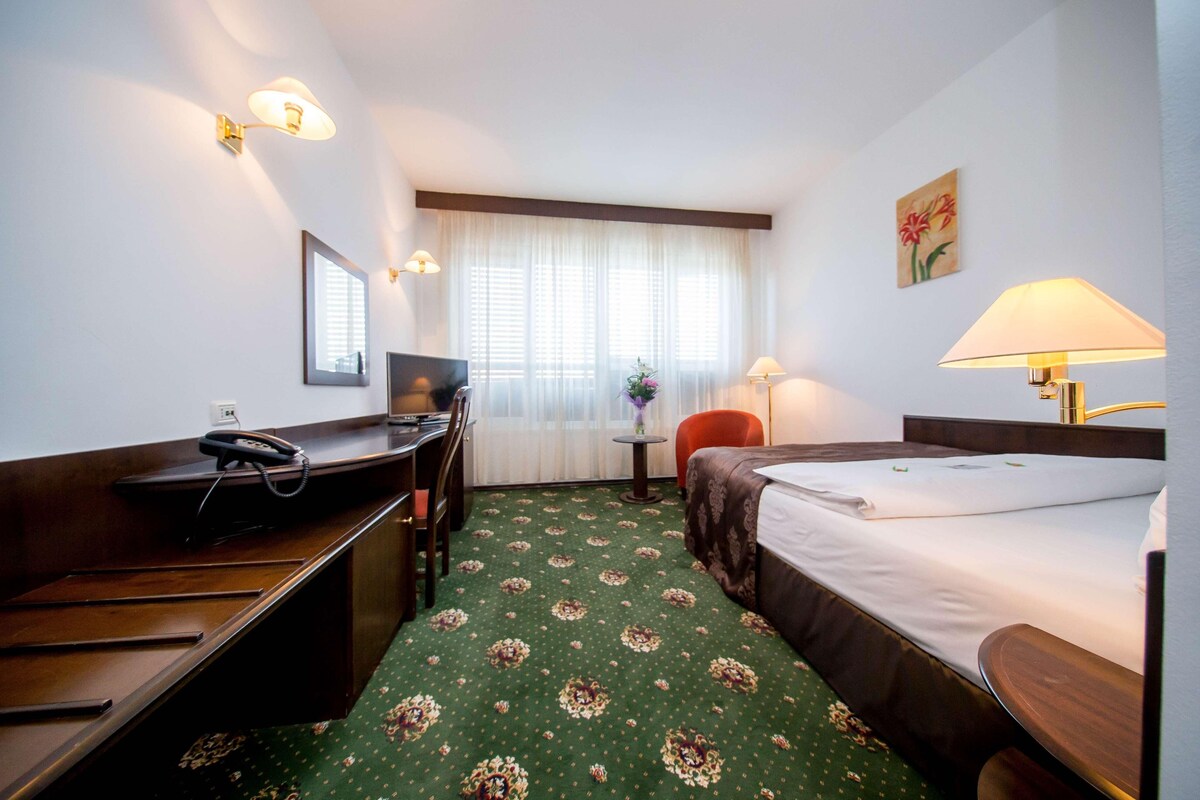 Habitación de hotel con escritorio, silla, cama y una mesita de noche con lámpara.