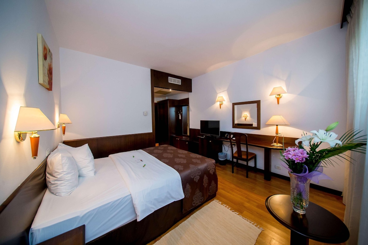 Una habitación de hotel con cama, escritorio, televisión y un florero con flores.
