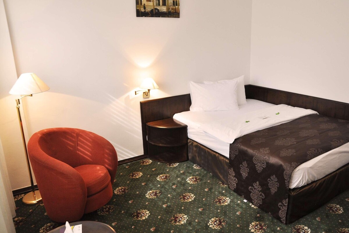 A hotel room with a single bed, a red chair, two bedside lamps, and a painting on the wall.