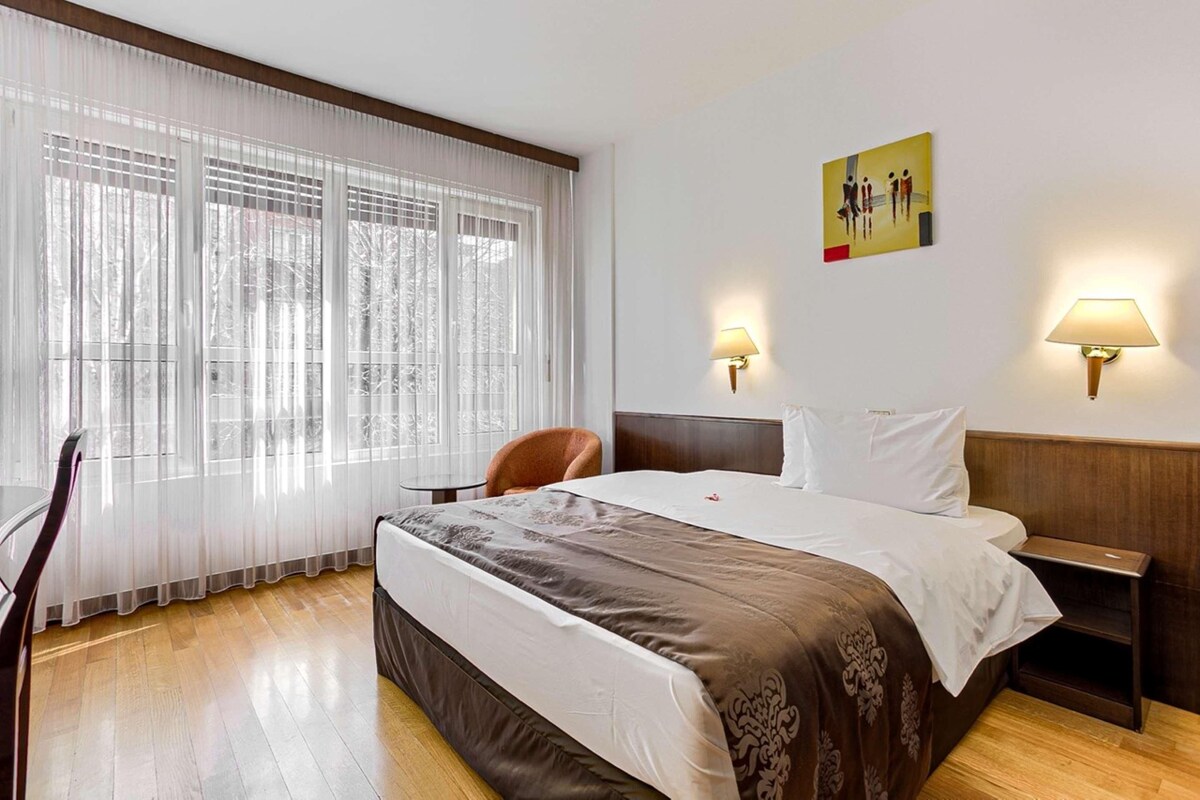 A hotel room with a bed, two bedside lamps, a chair, a small table, and a painting on the wall.