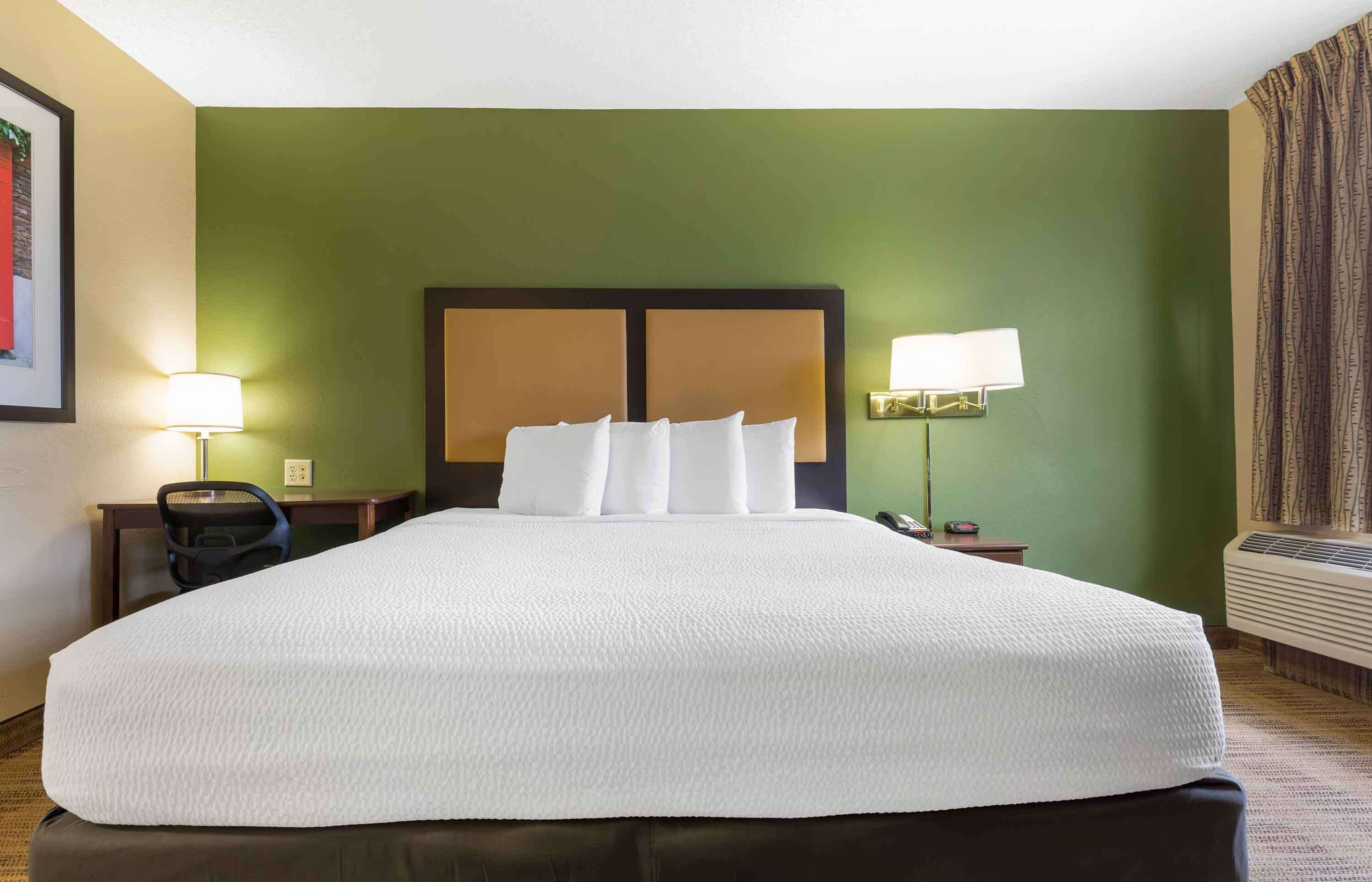 A hotel room with a large bed, two bedside lamps, a desk with a chair, and an air conditioning unit.