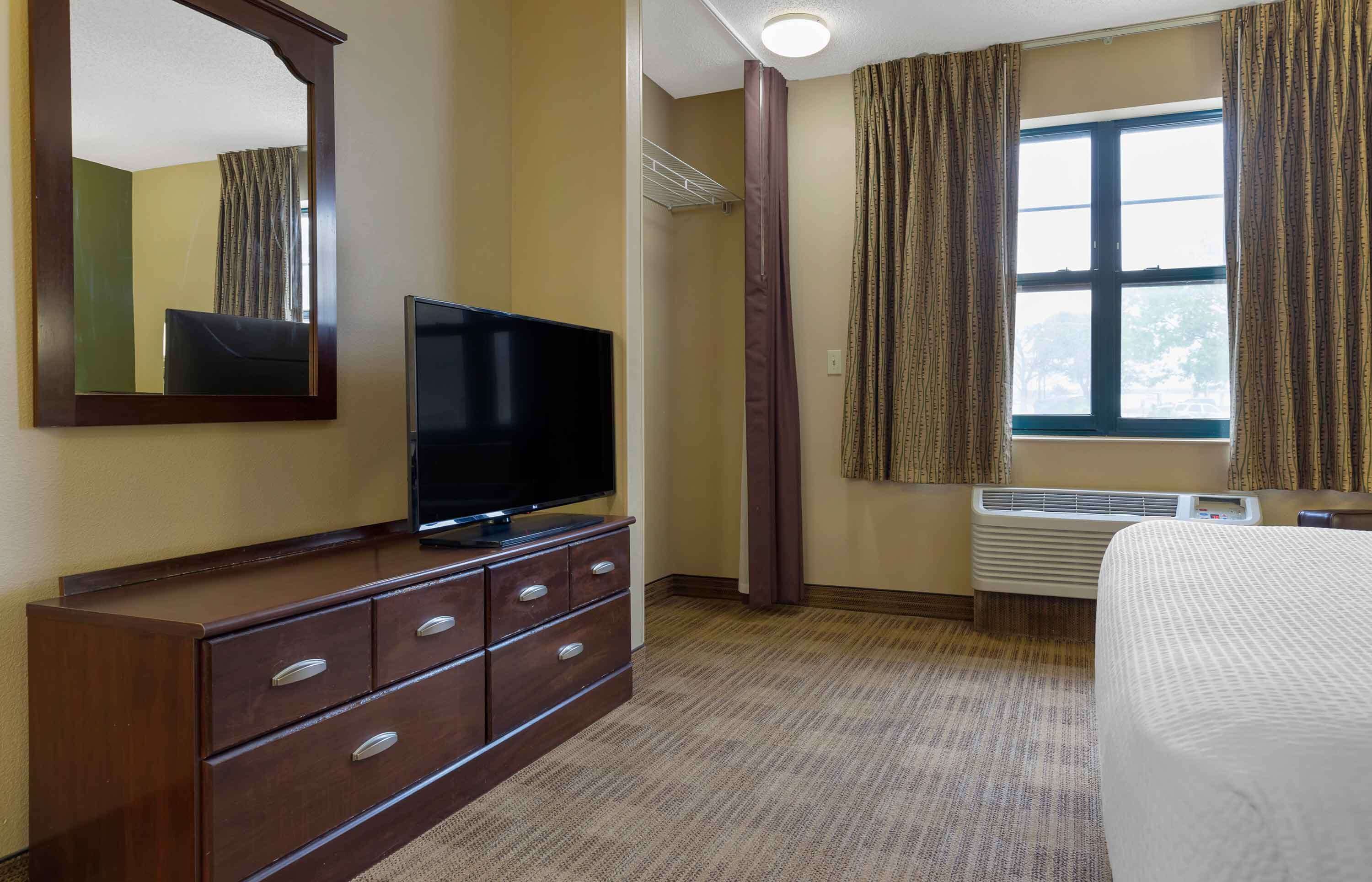 A hotel room with a flat-screen TV on a wooden dresser, a bed with a white cover, a window with curtains, and an air conditioning unit.