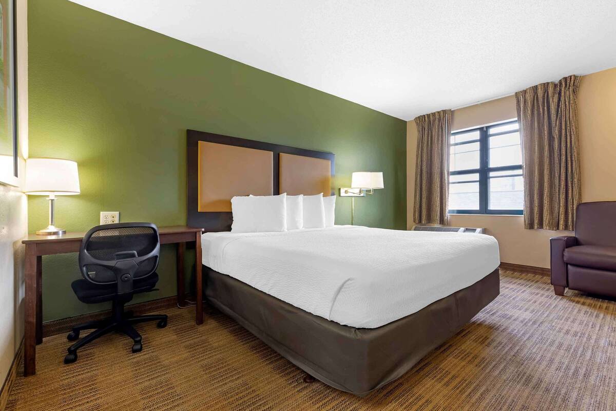 A hotel room with a large bed, a desk with a chair, a lamp, and a window with curtains.