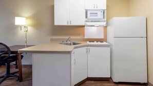 Cocina compacta con armarios blancos, microondas, refrigerador, pileta y una silla.