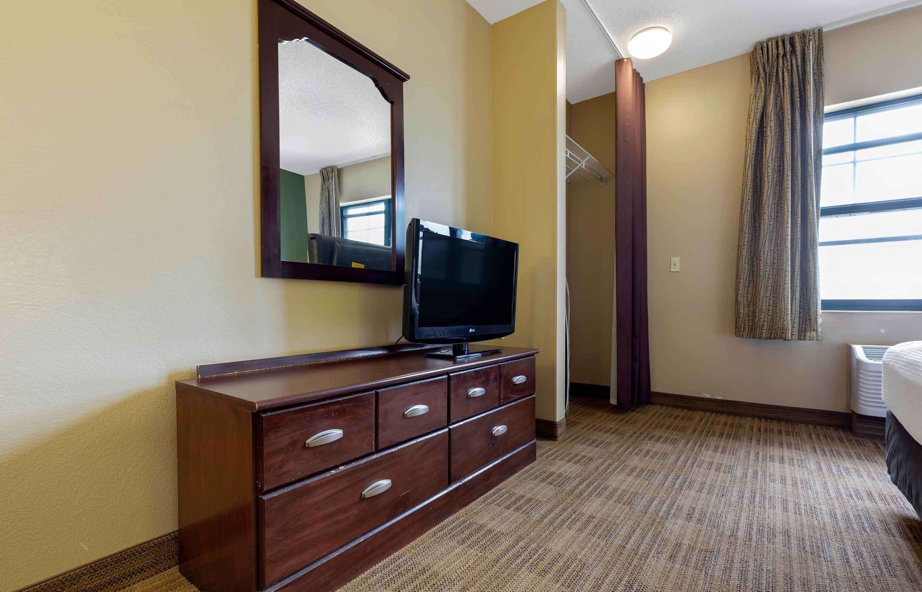 Quarto de hotel com cômoda de madeira, televisão de tela plana, espelho grande, janela com cortinas e piso coberto por carpete.