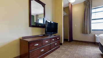 A hotel room with a wooden dresser, a flat-screen TV, a large mirror, a window with curtains, and a carpeted floor.