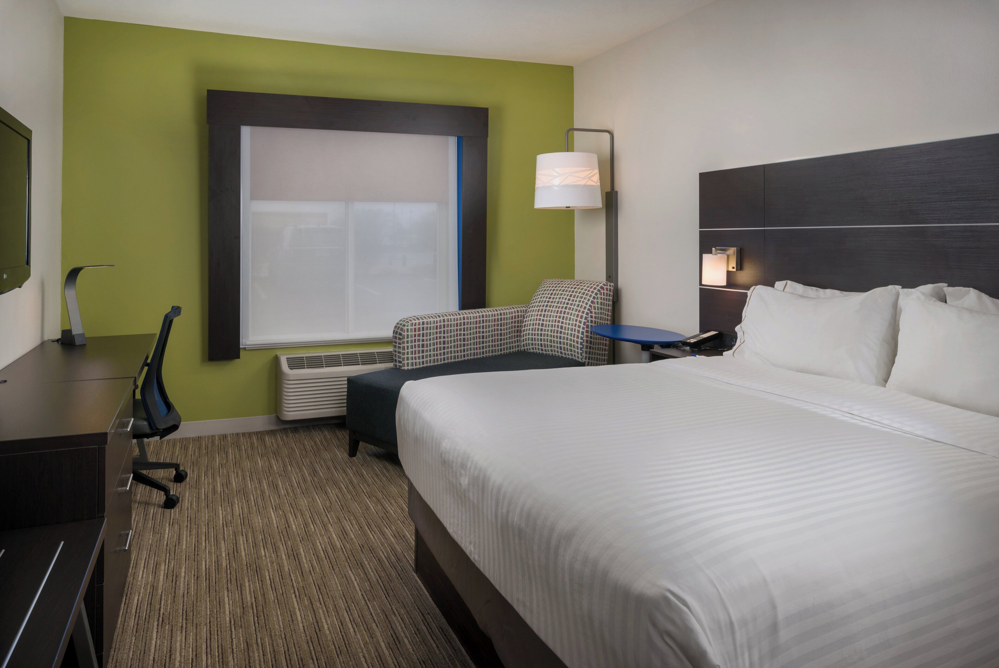 A hotel room with a bed, a desk, a chair, a lamp, and a window.